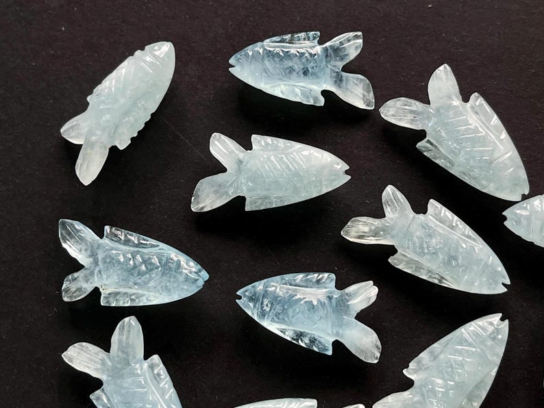 May include: A collection of light blue, carved gemstone fish figurines. Each fish has detailed fins and a carved body, with a slightly translucent appearance. The figurines are arranged on a dark surface.