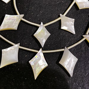 May include: Strands of iridescent, diamond-shaped mother-of-pearl beads. The beads are strung on a thin, silver-colored wire. The beads have a pearly white color with hints of yellow and iridescent shimmer. The beads are arranged in a repeating pattern.