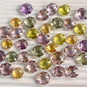 May include: A collection of 25 faceted star-shaped gemstones in shades of purple, pink, yellow, and green. The gemstones are arranged in a random pattern on a white surface.