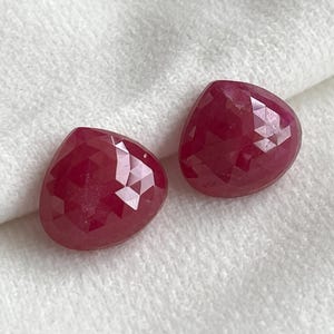 Pode incluir: Duas pedras preciosas soltas em forma de gota, de cor vermelho profundo e com uma superfície facetada. Cada pedra apresenta um acabamento polido com múltiplas facetas triangulares que refletem a luz, criando um padrão geométrico no topo.