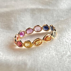Puede incluir: Un anillo de oro con una serie de piedras preciosas de colores. Las piedras son de forma ovalada y están dispuestas en un patrón continuo alrededor del anillo. Los colores incluyen rosa, morado, amarillo, naranja y azul. El anillo está sobre un fondo blanco texturizado.