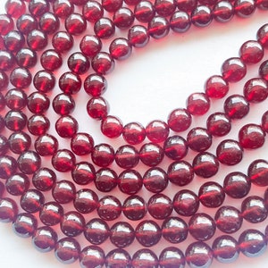 Könnte beinhalten: Nahaufnahme von mehreren Strängen tiefroter Edelsteinperlen. Die Perlen sind rund, poliert und haben eine glänzende Oberfläche. Sie sind aufgefädelt, möglicherweise für eine Halskette oder ein Armband. Die Perlen haben eine satte, dunkelrote Farbe.