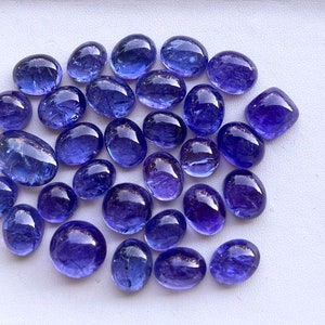 Puede incluir: Colecci&oacute;n de gemas de tanzanita pulidas, de forma ovalada y redondeada, de varios tama&ntilde;os. Las gemas muestran un color azul-violeta intenso y una superficie lisa y reflectante. Est&aacute;n dispuestas sobre un fondo blanco.