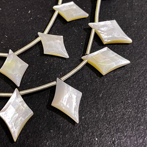May include: Strands of iridescent, diamond-shaped mother-of-pearl beads. Each bead is strung on a thin, silver-colored wire. The beads have a pearlescent sheen with hints of yellow and blue, and are set against a dark background.