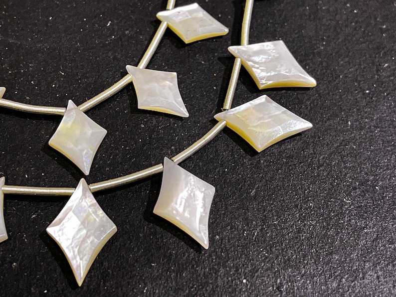 May include: Strands of iridescent, diamond-shaped mother-of-pearl beads. Each bead is strung on a thin, silver-colored wire. The beads have a pearlescent sheen with hints of yellow and blue, and are set against a dark background.