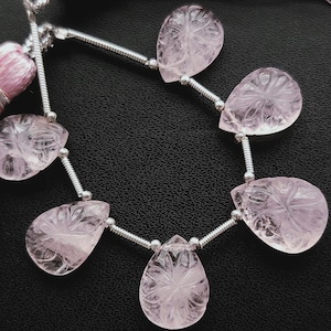 Puede incluir: Un collar de gemas rosas claras en forma de lágrima con intrincados diseños florales tallados. Las gemas están ensartadas con alambre plateado y pequeñas cuentas. Una borla rosa es visible en el lado izquierdo.