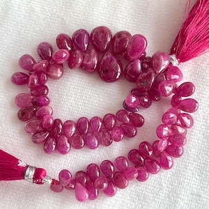 May include: A strand of teardrop-shaped, deep pink ruby beads arranged in a circular loop. The strand is secured at both ends with vibrant magenta tassels, each featuring a small silver-coloured metal band at the base of the thread bundle.