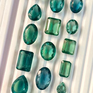 Può includere: Una collezione di gemme verdi e blu di varie forme e dimensioni, tra cui ovali, rotonde, a goccia e rettangolari, esposte su una superficie bianca.