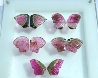 May include: Six carved gemstone butterfly wing pairs in shades of pink, green, and black. The wings are displayed on a white surface. The gemstones have a polished finish and are intricately carved with wing details.