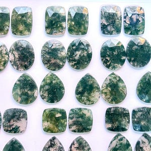 Op de afbeelding: Een verzameling van 20 mosagaten cabochons in verschillende vormen en maten. De stenen zijn een mix van groen, wit en bruin, met een draaiende structuur.