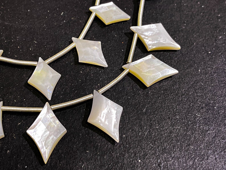 May include: Strands of iridescent, diamond-shaped mother-of-pearl beads. Each bead is strung on a thin, silver-colored wire. The beads have a pearlescent sheen with hints of yellow and blue, and are set against a dark background.