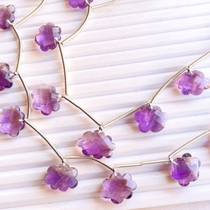 Peut inclure: Gros plan sur des brins de perles d'améthyste en forme de fleur. Chaque perle est d'un violet profond avec une coupe à facettes multiples, enfilée sur un fil fin de couleur argentée. Les perles sont posées sur un fond blanc strié.