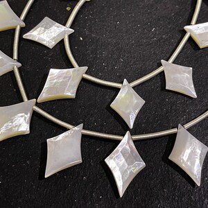 May include: Strands of iridescent, diamond-shaped mother-of-pearl beads strung on silver wire. The beads have a pearlescent sheen, reflecting light with a subtle rainbow effect. The beads are arranged in a repeating pattern.