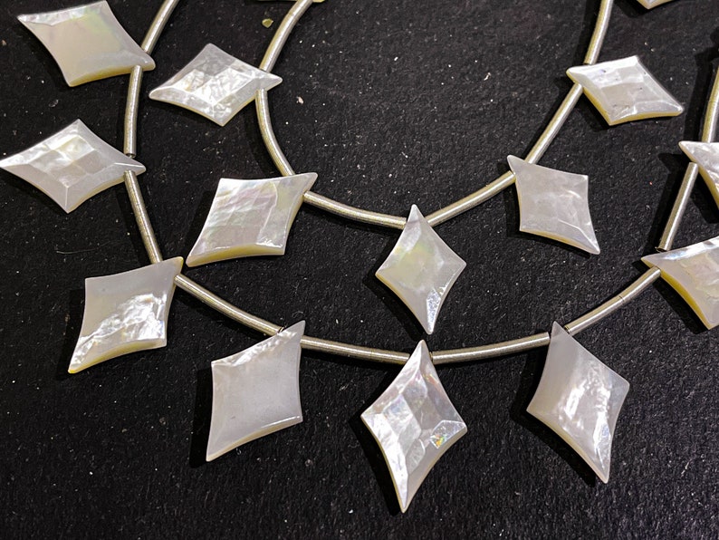 May include: Strands of iridescent, diamond-shaped mother-of-pearl beads strung on silver wire. The beads have a pearlescent sheen, reflecting light with a subtle rainbow effect. The beads are arranged in a repeating pattern.