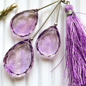 May include: Three purple amethyst teardrop shaped pendants with intricate carved floral designs, hanging from silver chains with a purple tassel.