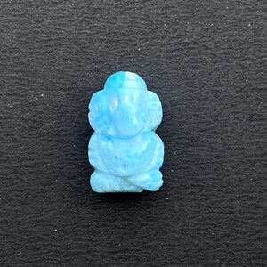 K&ouml;nnte beinhalten: Eine hellblaue, geschnitzte Figur einer sitzenden Gottheit, m&ouml;glicherweise Ganesha, mit gro&szlig;en Ohren und einem R&uuml;ssel. Die Figur besteht aus hellblauem Stein und ist vor einem dunklen Hintergrund platziert.