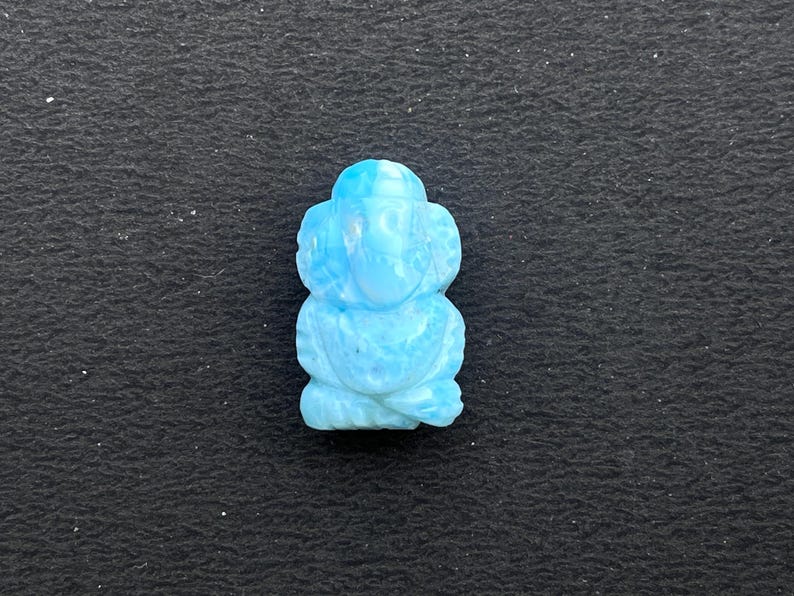 K&ouml;nnte beinhalten: Eine hellblaue, geschnitzte Figur einer sitzenden Gottheit, m&ouml;glicherweise Ganesha, mit gro&szlig;en Ohren und einem R&uuml;ssel. Die Figur besteht aus hellblauem Stein und ist vor einem dunklen Hintergrund platziert.