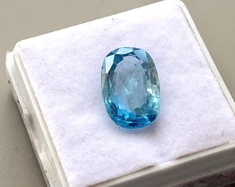 Könnte beinhalten: Ein ovaler, blauer Edelstein mit facettiertem Schliff. Der Edelstein wird in einer weißen Schachtel präsentiert.