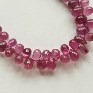 May include: A string of pink gemstone beads. The beads are teardrop-shaped and vary in shades of pink, from light to deep magenta. The gemstones are strung together, creating a necklace or bracelet.