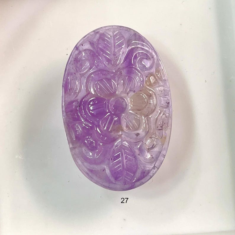 Puede incluir: Una piedra preciosa de amatista tallada con un dise&ntilde;o floral. La piedra preciosa es de color p&uacute;rpura claro y tiene un acabado liso y pulido.