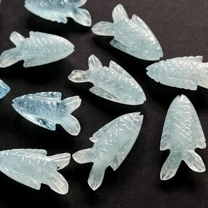 May include: A collection of aquamarine-colored, carved fish figurines. Each fish has detailed fins and scales, with a light blue hue. The figurines are scattered on a dark surface, creating a contrast that highlights their delicate features.