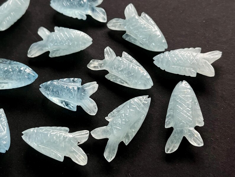 May include: A collection of aquamarine-colored, carved fish figurines. Each fish has detailed fins and scales, with a light blue hue. The figurines are scattered on a dark surface, creating a contrast that highlights their delicate features.
