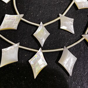 May include: Strands of iridescent, diamond-shaped mother-of-pearl beads. The beads are strung on a thin, silver-colored wire. The beads have a pearly white color with hints of yellow and iridescent shimmer. The beads are arranged in a repeating pattern.