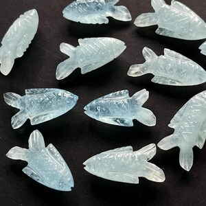 May include: A collection of light blue, carved fish figurines. Each fish has detailed fins and scales, and they are arranged on a dark surface. The translucent material gives them a delicate appearance, suggesting they are made of a gemstone or glass.