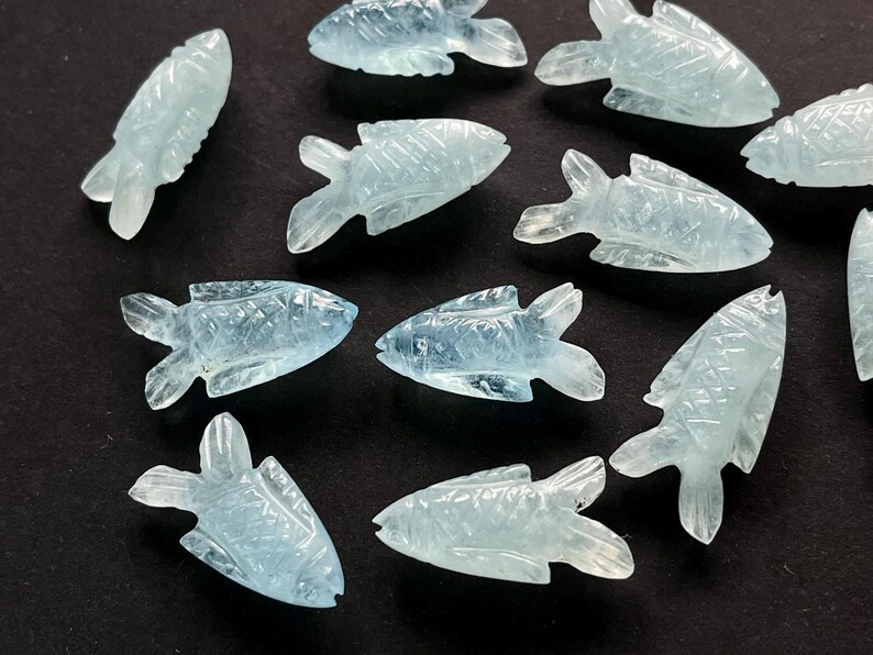 May include: A collection of light blue, carved fish figurines. Each fish has detailed fins and scales, and they are arranged on a dark surface. The translucent material gives them a delicate appearance, suggesting they are made of a gemstone or glass.