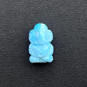 K&ouml;nnte beinhalten: Eine hellblaue, geschnitzte Figur einer Gottheit, m&ouml;glicherweise Ganesha, mit detailliertem Design. Die Figur ist vor einem dunklen, strukturierten Hintergrund platziert, der ihre Form und Farbe hervorhebt. Die Skulptur ist eine Darstellung einer religi&ouml;sen Figur.