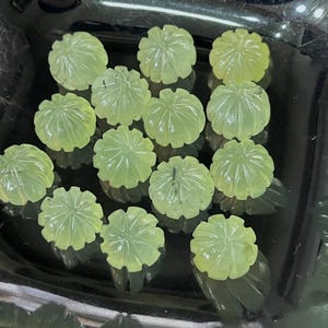 Puede incluir: Una colección de cuentas de piedras preciosas talladas de color verde claro. Cada cuenta tiene la forma de una flor estilizada con tallas detalladas de pétalos. Las cuentas se exhiben en un plato negro, mostrando su calidad translúcida y su intrincado diseño.