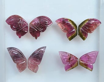 May include: Four pairs of carved gemstone butterfly wings in shades of pink, purple, and green. The wings are intricately detailed with carved lines and varying color gradients, showcasing the natural beauty of the stones. The wings are displayed on a white surface.