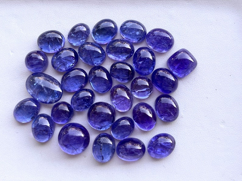 Puede incluir: Una colecci&oacute;n de piedras preciosas de tanzanita pulidas, de forma ovalada, en varios tama&ntilde;os. Las piedras muestran un color azul-violeta profundo con un acabado brillante. Las piedras preciosas est&aacute;n dispuestas sobre una superficie blanca, mostrando su belleza natural.