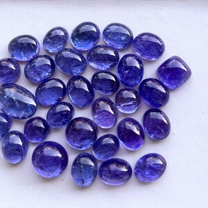 Puede incluir: Colecci&oacute;n de gemas de tanzanita pulidas, de forma ovalada y redondeada, de varios tama&ntilde;os. Las gemas muestran un color azul-violeta intenso y una superficie lisa y reflectante. Est&aacute;n dispuestas sobre un fondo blanco.