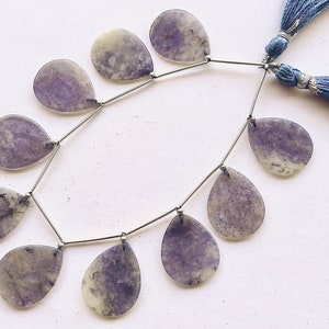 Könnte beinhalten: Eine Kette aus violetten und weißen Edelsteinperlen, auf einer silbernen Kette mit silbernen Perlenakzenten aufgefädelt. Die Perlen sind tropfenförmig und haben eine natürliche, polierte Oberfläche. Die Kette ist mit zwei blauen Quasten verziert.