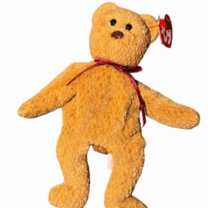 Peut inclure: Un ours en peluche brun doré avec un ruban rouge autour du cou. L'ours a de petits yeux noirs et un nez marron. Une étiquette rouge est attachée à son oreille. L'ours est debout, les bras et les jambes écartés.
