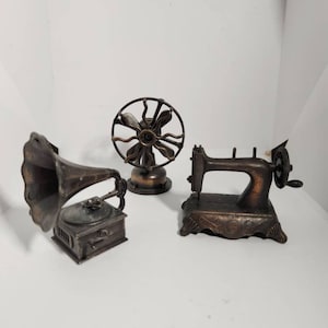Pode incluir: Três apontadores de lápis de metal estilo vintage: um ventilador, uma máquina de costura e um gramofone. Cada apontador é feito de metal escuro com acabamento bronze.