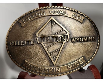 May include: A gold-colored metal belt buckle with the text "Triton Coal Co. Gillette Wyoming Buckskin Mine" and a diamond-shaped logo with the word "Triton" inside.