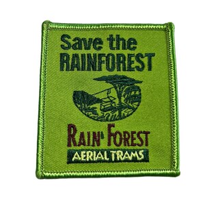 May include: Square green patch with a stitched border. The patch features the text "Save the Rainforest" in dark green, with an illustration of a rainforest and aerial trams. The text "Rain Forest Aerial Trams" is also displayed.