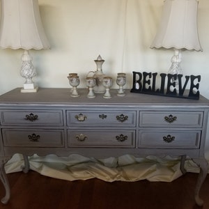 May include: A gray painted dresser with six drawers and carved legs. The dresser has black metal drawer pulls. Two lamps with white shades and a decorative pitcher and goblets are on top of the dresser. The word "BELIEVE" is displayed in black letters on the dresser.