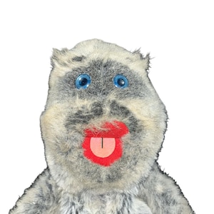 May include: A plush toy with a gray and white fur-like texture. It has large blue eyes, a red mouth, and a pink tongue. The toy's design is cartoonish and whimsical, with a friendly expression.