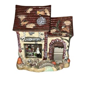 May include: A ceramic Halloween house with a red roof and a sign that reads "Laboratory". The house has a window with a scene of a witch and a ghost inside. There are skulls and pumpkins around the base of the house.