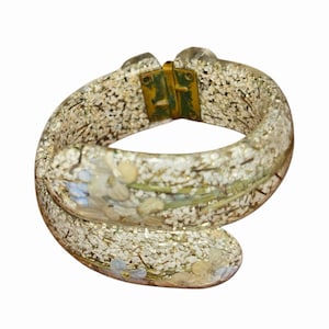 May include: A clear, resin bangle bracelet with embedded white, blue, and yellow floral elements. The bracelet has a gold-colored clasp and is filled with small, glittery particles. The overall design is a vintage, floral theme.