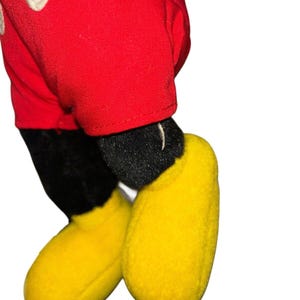 K&ouml;nnte beinhalten: Ein rotes und schwarzes Pl&uuml;sch-Mickey-Mouse-Spielzeug mit gelben Schuhen. Das Spielzeug steht auf einem wei&szlig;en Hintergrund.