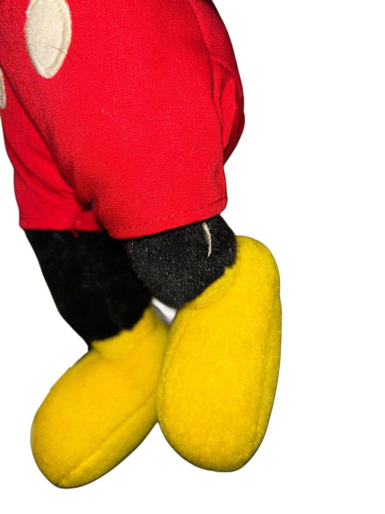 K&ouml;nnte beinhalten: Ein rotes und schwarzes Pl&uuml;sch-Mickey-Mouse-Spielzeug mit gelben Schuhen. Das Spielzeug steht auf einem wei&szlig;en Hintergrund.