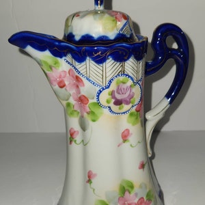 Puede incluir: Una tetera de porcelana blanca con un diseño floral azul y dorado. La tetera tiene una tapa y un asa. La tetera está decorada con flores rosas y moradas y hojas verdes.