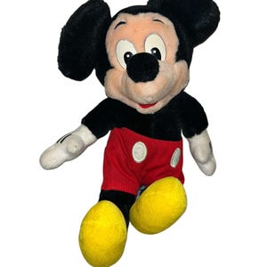 K&ouml;nnte beinhalten: Ein Pl&uuml;sch-Mickey-Mouse-Spielzeug mit schwarzen Ohren, einer schwarzen Nase und einem rot-wei&szlig;en Outfit. Das Spielzeug sitzt mit gekreuzten Beinen und hat ein gelbes L&auml;cheln.