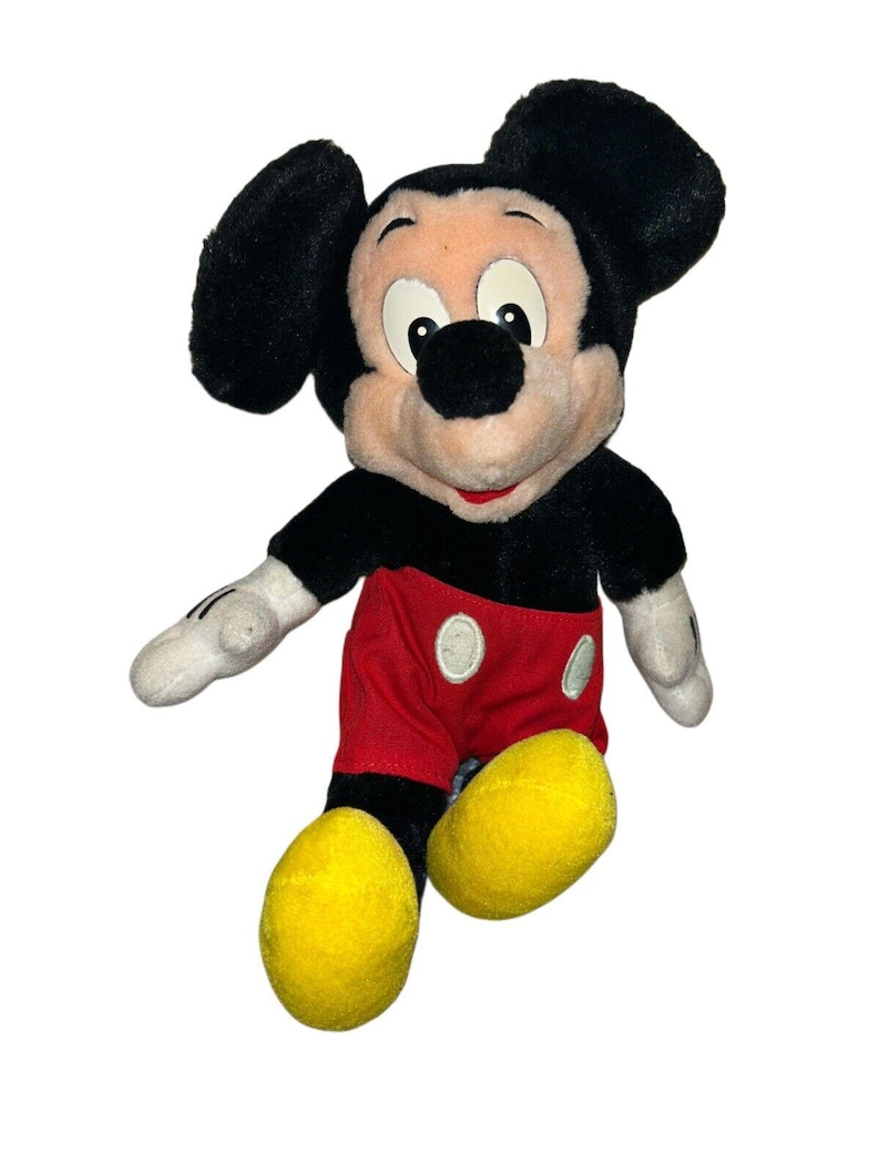 K&ouml;nnte beinhalten: Ein Pl&uuml;sch-Mickey-Mouse-Spielzeug mit schwarzen Ohren, einer schwarzen Nase und einem rot-wei&szlig;en Outfit. Das Spielzeug sitzt mit gekreuzten Beinen und hat ein gelbes L&auml;cheln.