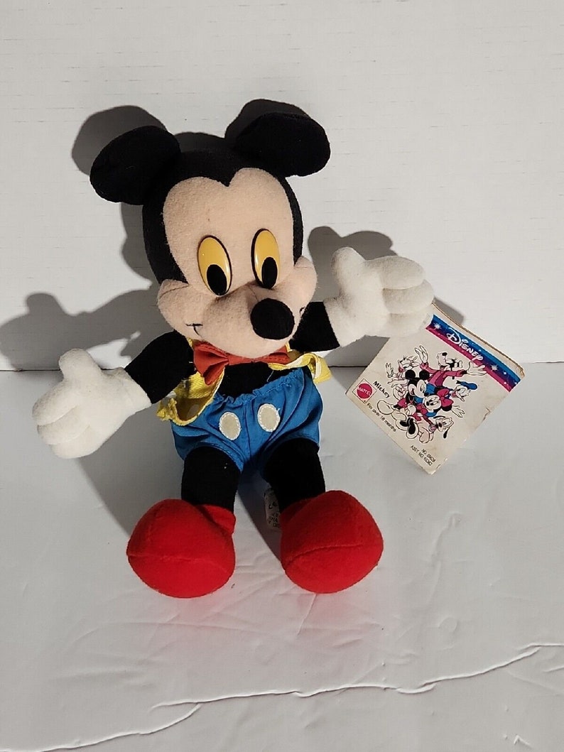 K&ouml;nnte beinhalten: Ein Pl&uuml;sch-Mickey-Mouse-Spielzeug, das ein gelbes Hemd, eine blaue Latzhose und eine rote Schleife tr&auml;gt. Das Spielzeug sitzt auf einer wei&szlig;en Oberfl&auml;che und hat ein Disney-Etikett am rechten Arm.