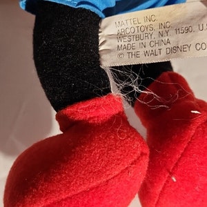 K&ouml;nnte beinhalten: Ein schwarzes und rotes Pl&uuml;schtier mit einem blauen Hemd. Das Spielzeug hat ein Etikett mit der Aufschrift "MATTEL INC. ARCO TOYS, INC. WESTBURY, N.Y. 11590, U.S. MADE IN CHINA &copy; THE WALT DISNEY CO."